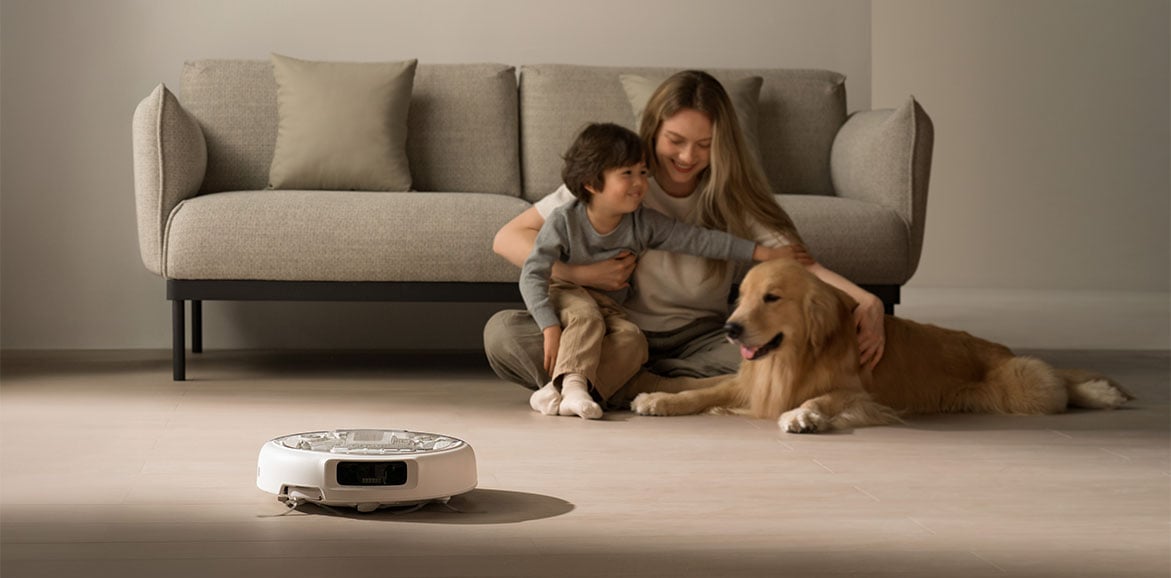 Robot sprzątający DJI ROMO A sprząta obok siedzącej na podłodze kobiety z dzieckiem i psem